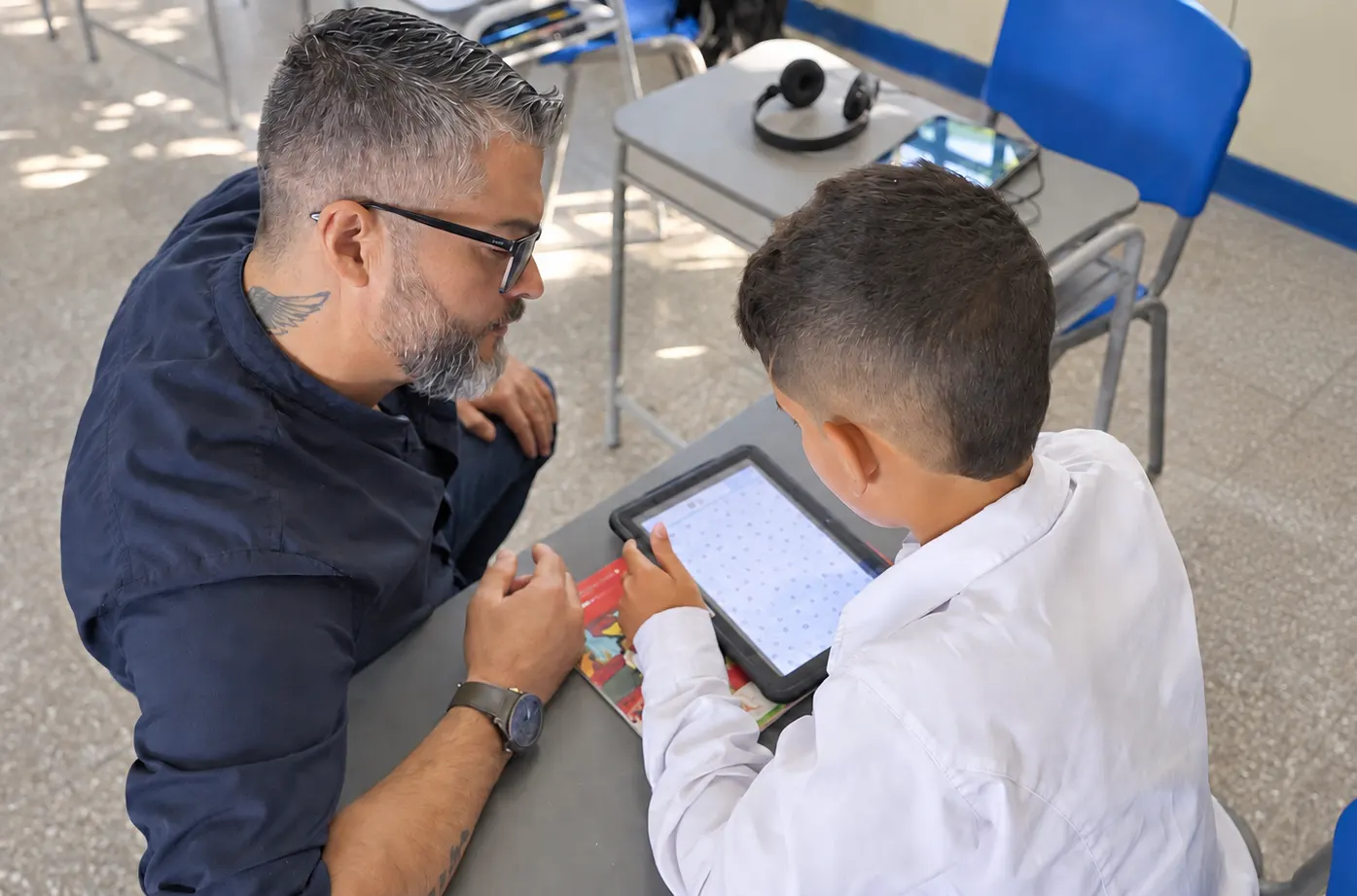 A teacher sitting next to a student follows an activity on a tablet with him; on the desk behind there are headphones and another tablet