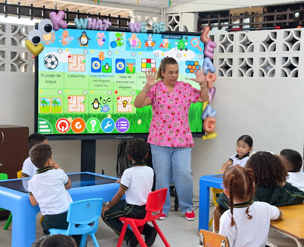 A teacher in front of an AVACOM interactive screen showing a menu of labelled activities, while the class watches from the interactive tables
