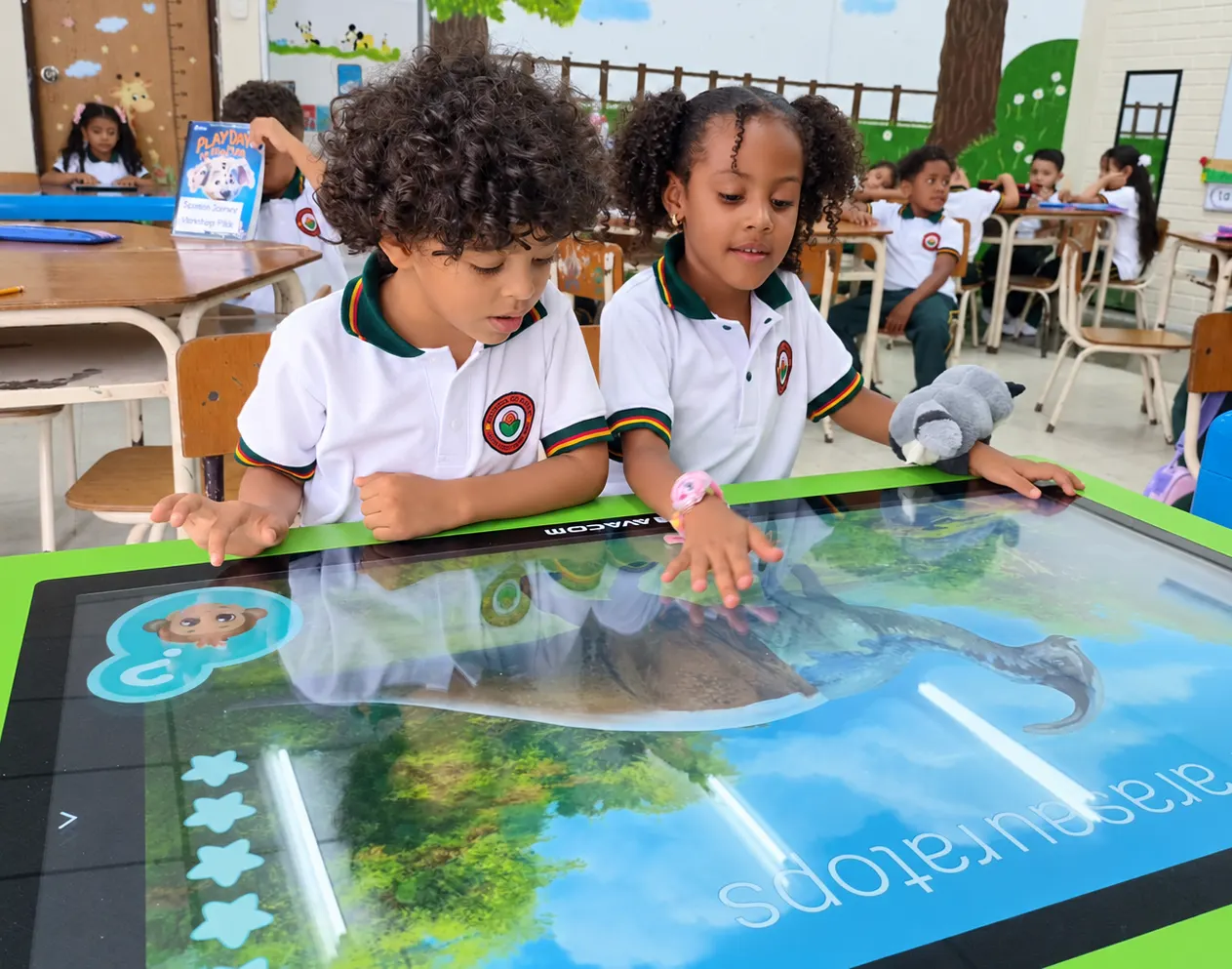Two children rest their hands on a green-framed AVACOM interactive table showing a dinosaur and its name