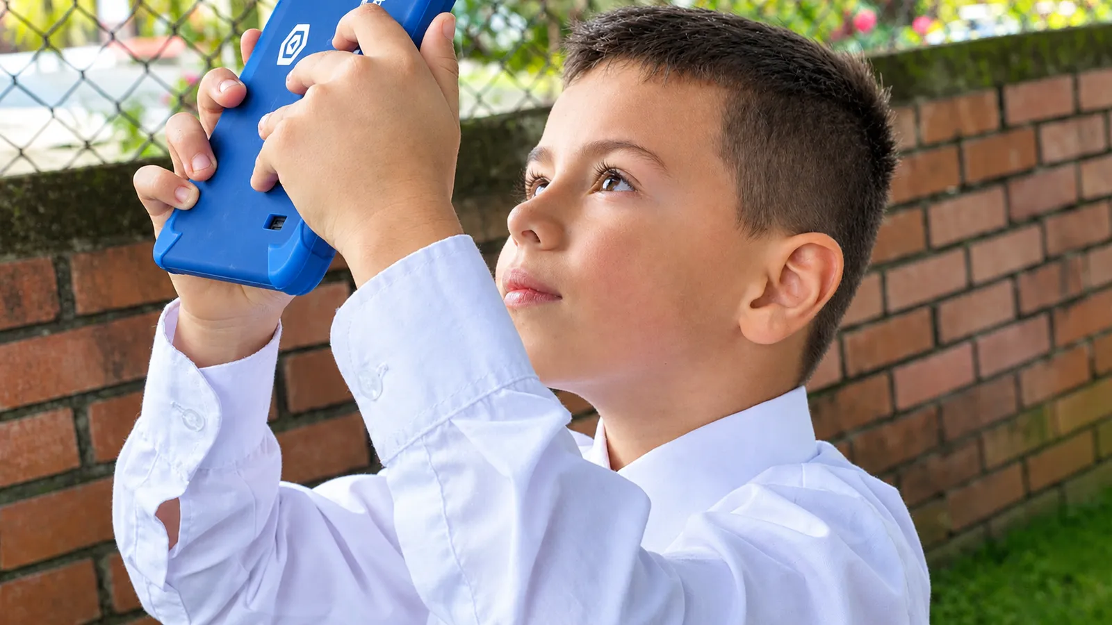 A boy in a white shirt holds a blue tablet up high and looks at it attentively, in front of a brick wall and a wire fence
